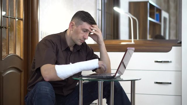 A man with a broken right hand in a plaster sits near a coffee table with a laptop in a room with furniture. Broken arm