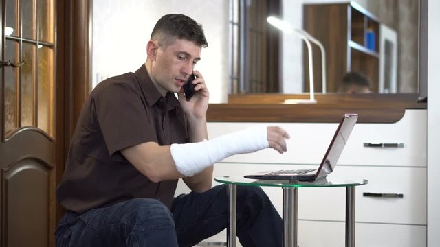 A man with a broken right hand in a plaster sits near a coffee table with a laptop and talks through a smartphone in a room with furniture. Broken arm