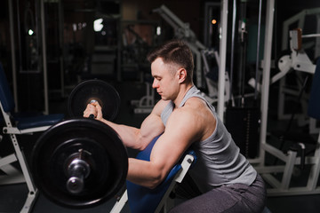 Fototapeta na wymiar Strong handsome fit man exercising in the gym. Personal trainer workout. Athletic man working out his arms muscles with barbell on a bench. Fitness, healhty lifestyle, bodybuilding concept.
