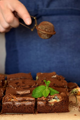 Brownie square pieces decorated with mint and a confectioner wearing navy blue apron is covering cakes with cacao powder