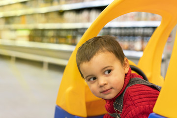 Fototapeta na wymiar Toddler boy looks out of a shopping cart for children in a supermarket on the background of shelves with food