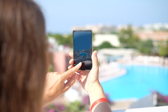 Happy woman taking photos of fantastic view with full view smartphone camera on summer travel vacation. Brunette girl with long hair standing backwards and making photography towards the swimming pool