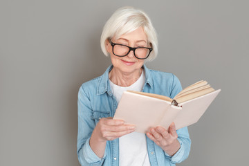 Senior woman in jeans jacket studio isolated on grey wall reading book joyful