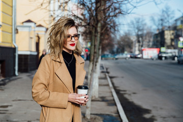 Fashion girl in a beige coat and a paper cup of coffee in her hands.