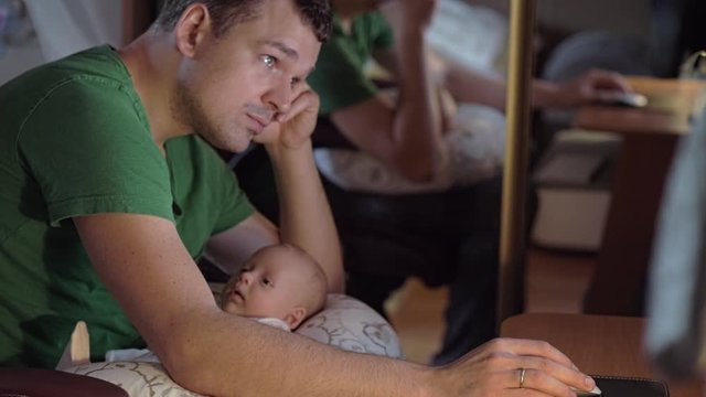 Tired dad working on the computer and looking after baby daughter lying on his lap. Family and job