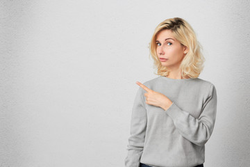 Naklejka premium uncertain young female with curly blonde hair indicates with finger aside at blank copy space. Serious expression. Isolated over white wall