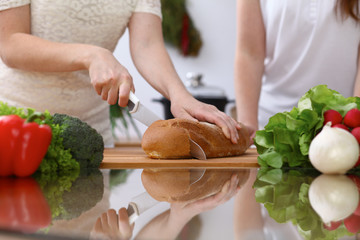 Closeup of human hands cooking in kitchen. Mother and daughter or two female cutting bread at the table full of vegetables and green salad. Healthy meal, vegetarian food and lifestyle concepts