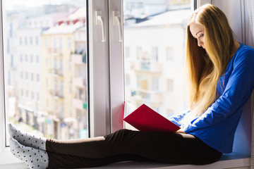 Woman sitting on window sill reading book at home