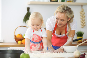Obraz na płótnie Canvas Little girl and her blonde mom in red aprons playing and laughing while kneading the dough in the kitchen. Homemade pastry for bread, pizza or bake cookies