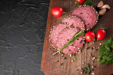 Smoked salami on a black table and a brown board. Sausages with rosemary, garlic and pepper and tomato cherry, dill, parsley.
