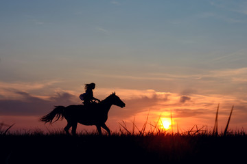 Fototapeta na wymiar Romantic equine and girls silhouette on horse hiking with red rising sun on horizon. Galloping horse with female rider on beautiful colorful sunset background. 
