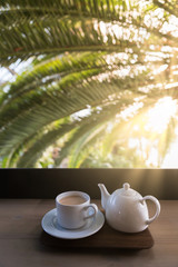 White teapot with cup of Masala tea on the table near a palm tree and wonderful light with sun rays, vertical