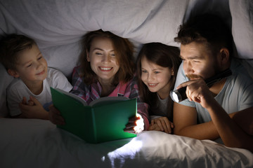 Happy family reading bedtime story under blanket in evening