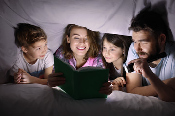 Happy family reading bedtime story under blanket in evening