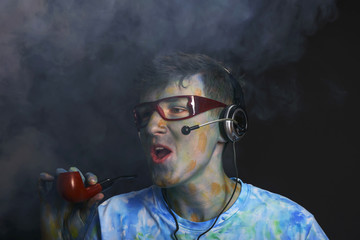 Young interesting guy painted with colorful paints in glasses and a headset smokes a pipe.
