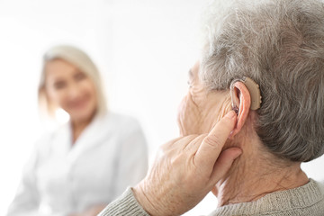 Senior woman adjusting hearing aid on light background, closeup
