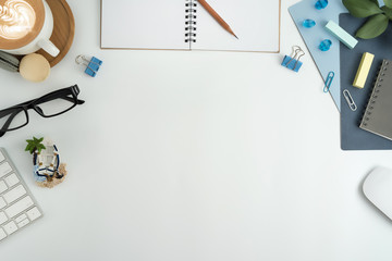 Flat lay, top view office table desk. Workspace with blank note book, keyboard, macaroon, office supplies, white flowers, green leaf, blue ornament and coffee cup on white background.