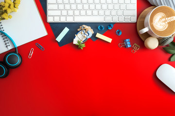 Flat lay, top view office table desk. Workspace with blank note book, keyboard, macaroon, office supplies, yellow flowers, green leaf, blue ornament and coffee cup on red background.