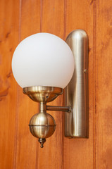 A house lamp with a brass body and a round white matte lampshade in the old English style hangs on the wall of wooden boards painted in brown