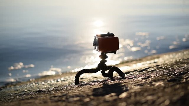 Concrete embankment with an action camera in waterproof plastic case with float on gorilla tripod filming blue water of sea, river or ocean with sunshine reflected in water