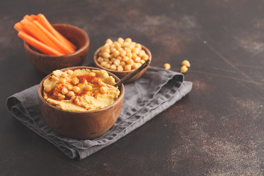 Hummus, fresh carrot sticks and boiled chickpeas in wooden bowls. Vegan food concept, dark background, copy space.