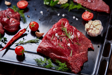 Flat lay of raw beefsteak with vegetables, herbs and spicies on metal tray, close-up, selective focus