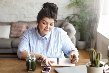 Hardworking positive young female freelancer with heavy curvy body multitasking at home office,...
