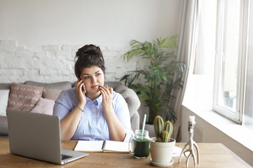 Horizontal shot of concentrated chubby young woman with hair bun multitasking at home office, sitting at desk in front of open laptop computer, making notes in diary and having phone conversation - Powered by Adobe