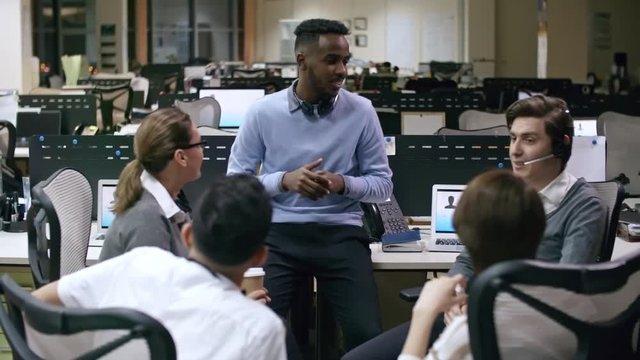 PAN of male African call centre agent sitting on desk and chatting with group of colleagues during break