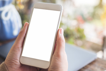 Mockup image of hands holding white mobile phone with blank desktop screen with laptop on table in cafe