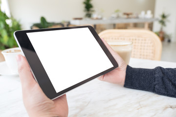 Mockup image of hands holding black tablet pc with blank white screen and coffee cup on table in cafe