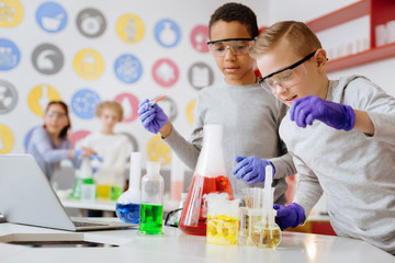 Experimenting with chemicals. Two upbeat teenage friends using pipette and adding some chemical agents to the flask and test tubes while having the chemistry class in the lab