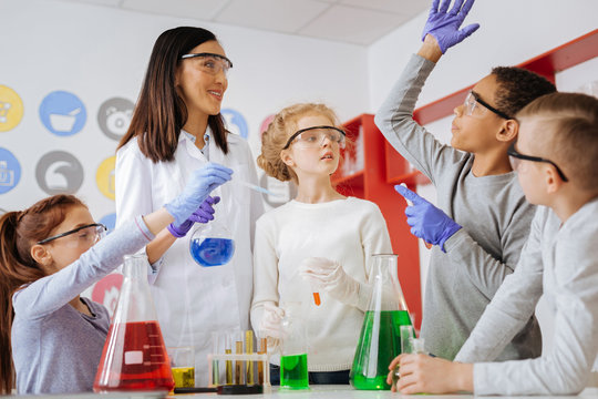 Important question. Pleasant teenage male student raising a hand and asking his teacher a question while she and his classmates conducting a common chemical experiment