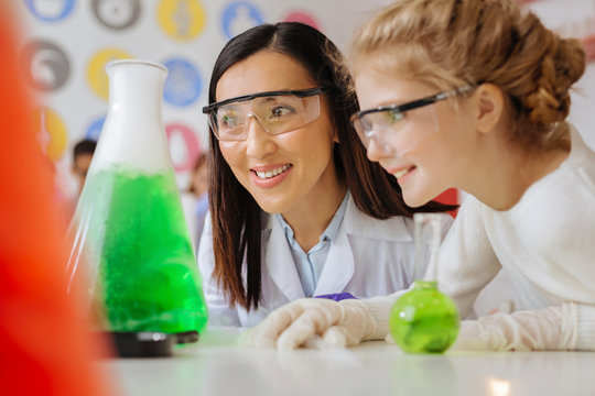 Perfect ambience. Cheerful teenage girl and her chemistry teacher sitting at the table in the lab and smiling pleasantly while watching a chemical reaction in the flask