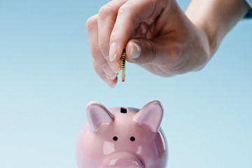 Partial view of woman putting coin into pink piggy bank isolated on blue