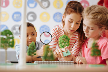 Close view. Pleasant teenage boy looking at a tree model through a magnifying glass while his classmates holding the model in the hands and scrutinizing it