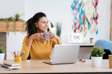 Good day. Beautiful exuberant young curly-haired woman smiling and drinking tea while working in the office