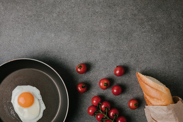 top view of fried egg on frying pan, fresh tomatoes and baguette in paper bag on grey