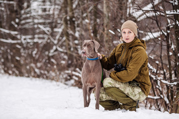 Female hunter in camouflage, armed with a rifle, standing in a snowy winter forest with duck prey