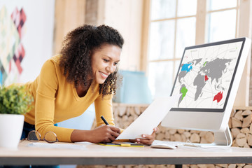 My office. Pretty cheerful young curly-haired woman holding and studying a sheet of paper and standing at her table