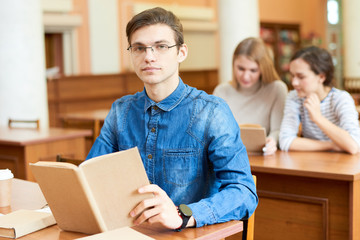 Serious confident handsome young male student preparing for graduation exam and reading textbook in modern library and looking at camera