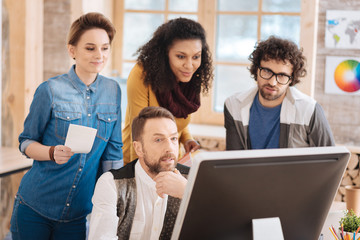 Fototapeta premium Getting project done. Handsome unsmiling bearded man working on his laptop and talking with his colleagues and sitting at the table