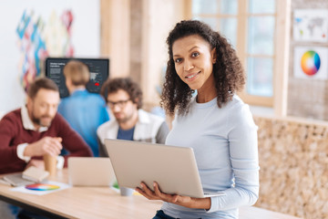 Best worker. Beautiful content curly-haired woman smiling and holding her laptop while her colleagues working in the background