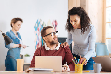 Fototapeta premium Delighted. Handsome content bearded man working on his laptop while sitting at the table and his colleague standing behind him