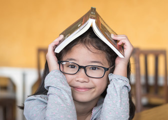 Educational literacy, world book day concept with smart Asian school student girl kid with eyeglasses having children book over her head, thinking of happiness in reading for young reader
