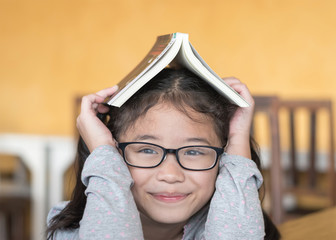 Educational literacy, world book day concept with smart Asian school student girl kid with eyeglasses having children book over her head, thinking of happiness in reading for young reader