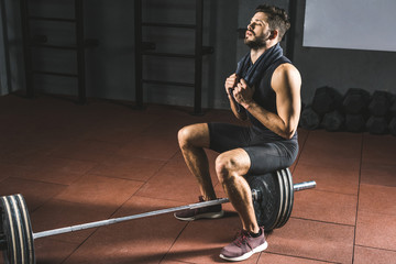 Fototapeta na wymiar Tired sportsman with towel sitting on barbell in gym