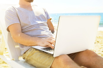 Fit young man sitting on deck chair at sea view beach using laptop. Male freelance programmer in chaise-long lounge working coding blogging surfing on notebook computer. Perks of distance work concept