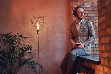 Portrait of a stylish man in a flannel suit and glasses listening music with the phone and...