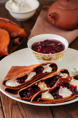 Chocolate crepes (blinis) with sour cream and black currant sauce on a white plate. Wooden background.